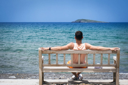 年轻男子伸出双臂, 坐在一个木凳上的地中海沿岸的克里特岛上, 并期待着地平线