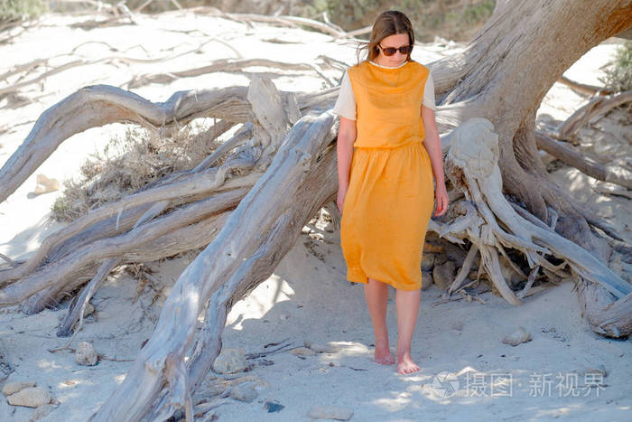穿裙子的年轻女孩站在附近的树上的白色沙滩上赤脚