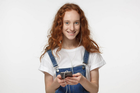 微笑卷曲红发女孩的肖像手持智能手机, 看着相机, 听音乐, 孤立在灰色背景