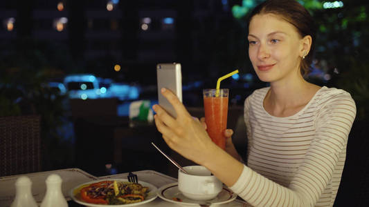幸福的女人拍拍照使用智能手机和 drinck 在晚上坐在餐厅里的鸡尾酒汁
