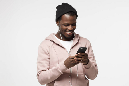 年轻英俊的黑人男子在粉红色的帽衫和黑色的帽子, 听音乐与耳机和看他的智能手机屏幕
