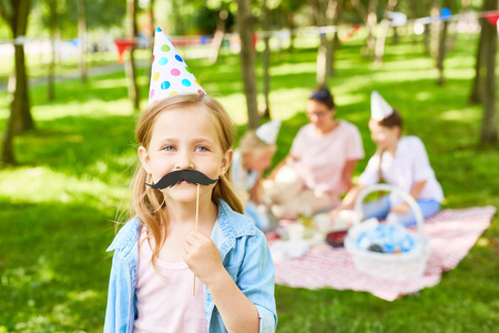 逗人喜爱的女孩在她的嘴举行滑稽的小胡子在室外生日党
