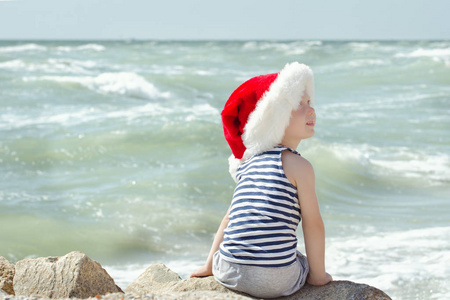 男孩在圣诞老人的帽子和条纹 tshirt 坐在海滩上。后视图