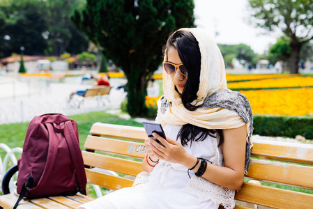 年轻的阿拉伯妇女的围巾，眼镜坐在板凳上，看起来她的智能手机