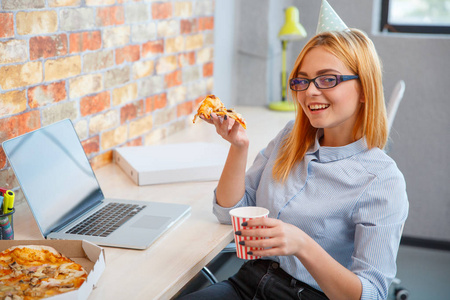 一个女孩在吃比萨饼在办公室内，面对一台笔记本电脑
