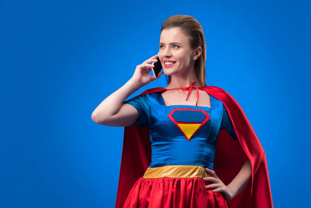 在超级英雄服装的微笑的妇女的画像谈论在蓝色被隔绝的智能手机
