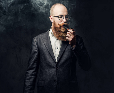 红发的大胡子男性身着西装和眼镜吸烟传统管道在深灰色背景照片