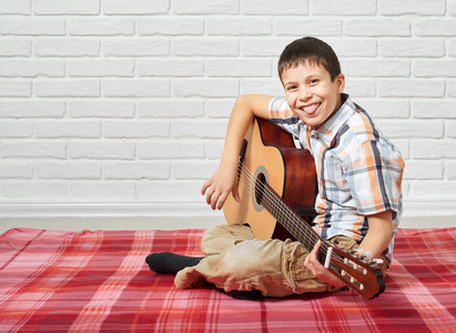 男孩在吉他演奏音乐, 坐在红色格子毯子, 白色砖墙背景