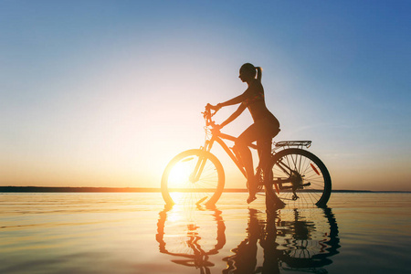 一个穿着五颜六色西装的金发碧眼的女人在温暖的夏日里, 在夕阳下的水中坐在自行车上。健身理念。天空背景