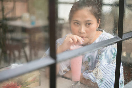 亚洲妇女在咖啡馆坐在玻璃墙壁在一个雨天和喝草莓牛奶