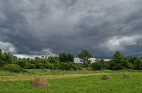 田野里有成堆的割草, 乡下的房子在背景和暴风雨的天空