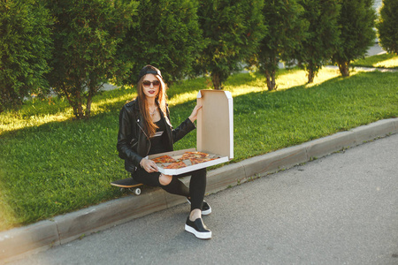 年轻的时髦女人与滑板有一个午餐与切片的比萨饼坐在户外
