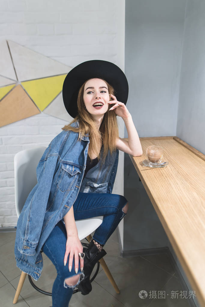 一个时髦的女人在一个舒适的咖啡馆里对着相机摆姿势, 在窗边喝杯热饮