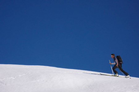 在晴朗的蓝天下, 男性背部滑雪者沿着雪脊往山顶上徒步。