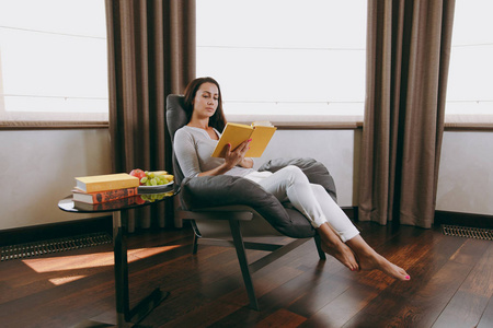 美丽的年轻女子在家坐在现代椅子在窗口前面, 放松在她的客厅和读书书
