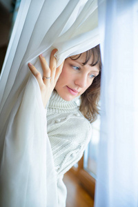 舒适家美丽的惊讶时尚的女人在一个家庭设置看着窗外拿着窗帘。女性肖像