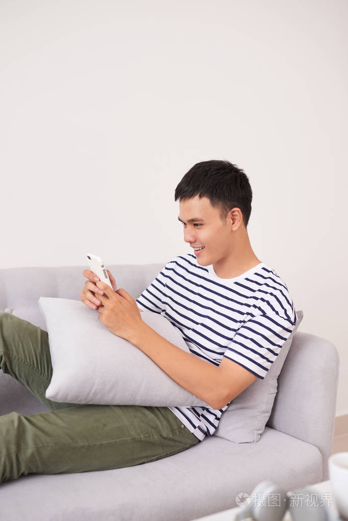 年轻的亚洲男子坐在沙发上放松智能手机检查他的智能手机信息