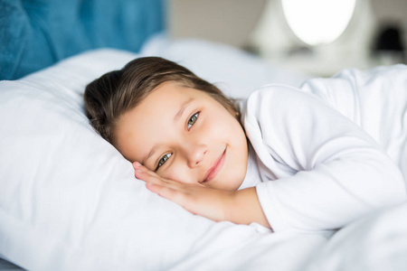 特写笑的小女孩躺在床上, 手放在枕头下, 看着相机和微笑。早上好