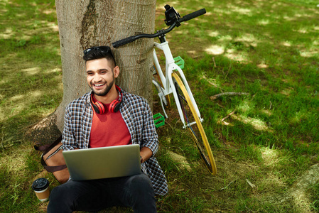 笑的年轻印度男子坐在树上, 他旁边的计算机和自行车