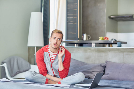 年轻的现代男子在 homewear 讲智能手机, 而坐在床上