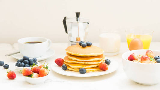 与蓝莓草莓和黑咖啡牛奶和橙汁在白色石头桌背景的健康早餐设置与薄饼和麦片