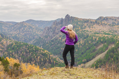 一个穿紫丁香夹克的女孩看着远处的山, 山的景色和一个阴天的秋天森林。文本的可用空间