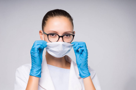 聪明的年轻女医生戴着眼镜和白色防护面具的脸, 戴着蓝色手套看着相机