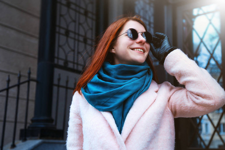 红头发美丽的女孩走在街道上的粉红色的大衣和蓝色的围巾, 与太阳镜