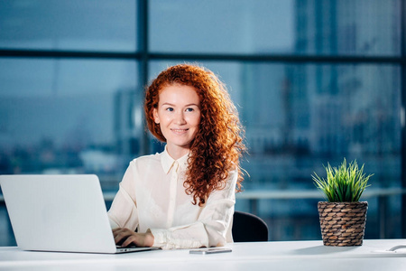 红发女实业家坐在明亮的现代工作站上, 在笔记本电脑上打字