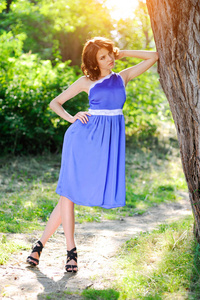 一个年轻的黑发女孩穿着蓝色的连衣裙, 在夏日公园的一棵大树旁, 在明亮的阳光下