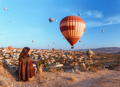 妇女在传统的雨披衣服观看美丽的景色, 五颜六色的热气球飞越山谷在土耳其。