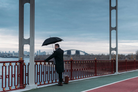 带雨伞和手提箱的时尚孤独男子站在桥上