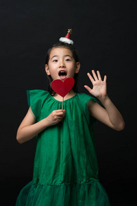 可爱的亚裔女孩穿着绿色的礼服, 拿着圣诞装饰品, 一根心贴在黑色的背景上。一张惊讶的脸