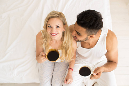 年轻夫妇喝咖啡坐在床上, 快乐的微笑拉美裔男人和女人的顶角视图