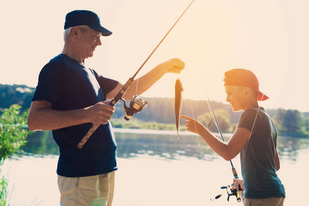 那位老人和他的孙子正站在河岸上。老人抓住鱼, 向男孩展示渔获物