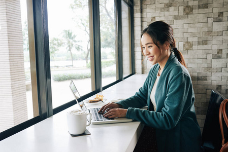 美丽的年轻亚洲妇女坐在窗口在咖啡馆和工作在膝上型电脑