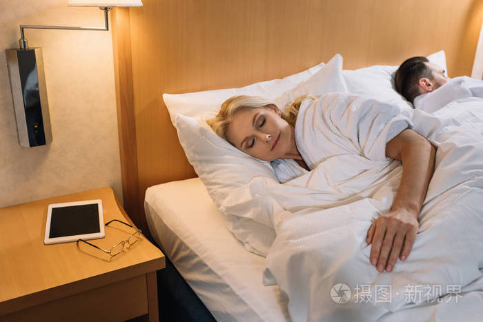 成熟夫妇在床上睡觉的浴袍和数字片剂与镜片在桌在旅馆房间