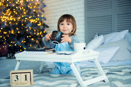 元旦前夕, 12月31日。可爱的小女孩在睡衣与杯子在圣诞节树附近