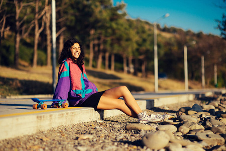 穿着紫色夹克的快乐时尚女孩休息, 坐在滑板旁边