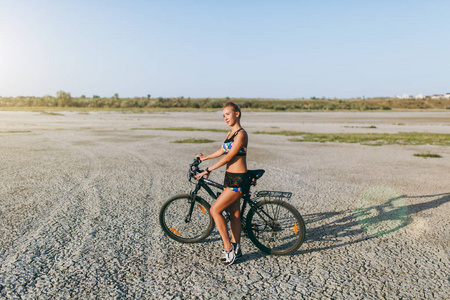 一个强壮的金发女郎穿着五彩斑斓的西装站在沙漠地区的自行车旁, 看着太阳。健身理念。蓝天背景