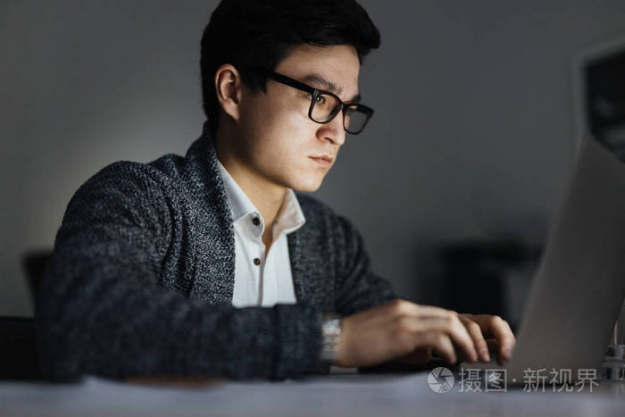 侧面图亚洲男子戴眼镜工作在黑暗的房间深夜, 他的脸被屏幕照亮