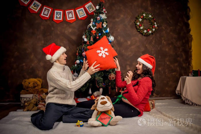 微笑美丽的未来父母是愉快地玩枕头, 而坐在地板附近的装饰圣诞树。水平射击