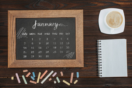 顶级的咖啡, 空的笔记本, 五颜六色的粉笔和黑板与1月日历的顶部视图