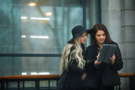 两个漂亮的女孩穿着暖和的衣服讨论在大楼附近的街道上的一块平板电脑工作