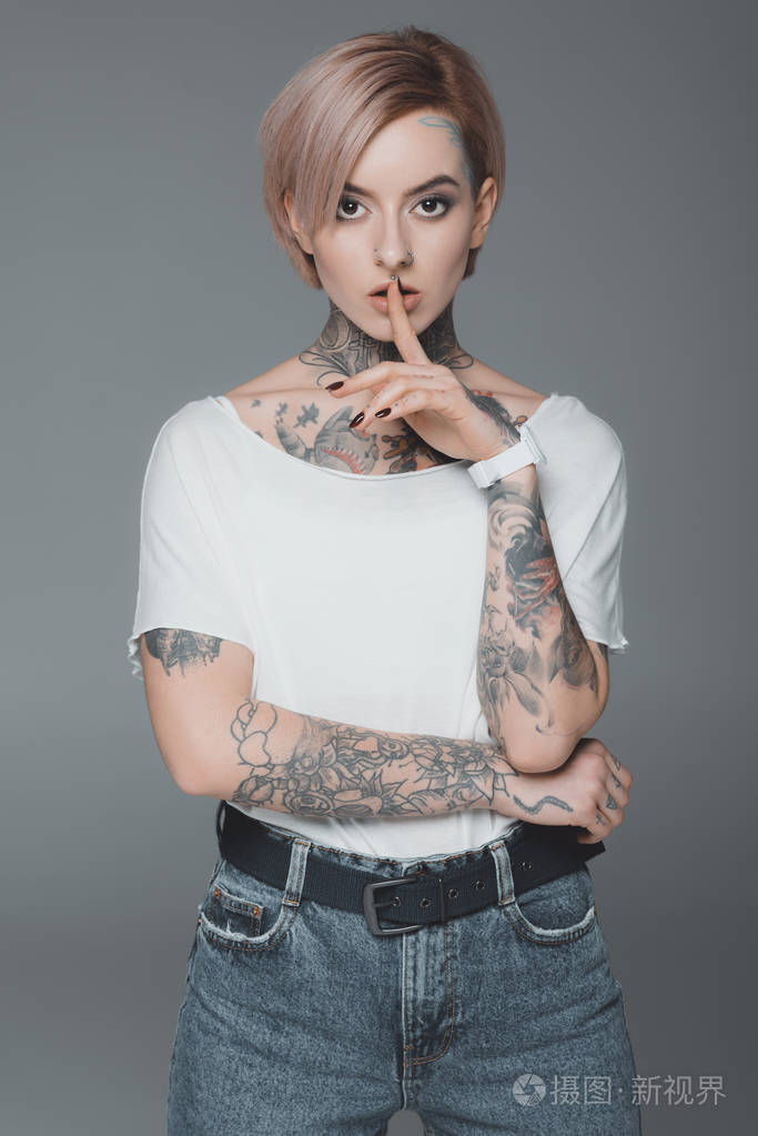 美丽的纹身女孩打手势沉默, 看着相机孤立在灰色