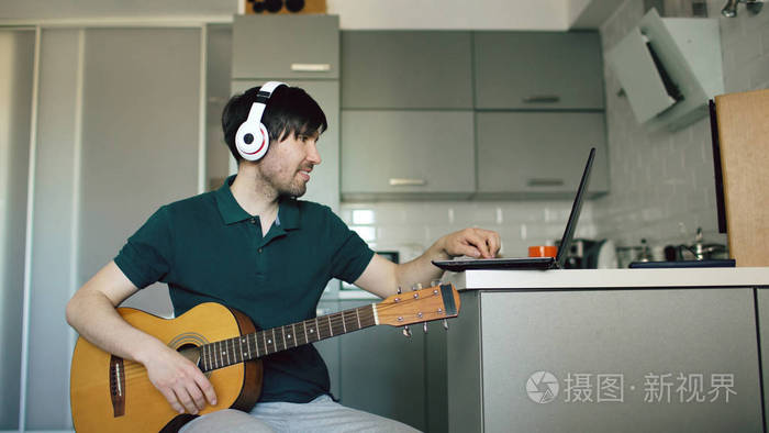 活泼开朗的年轻人戴着耳机坐在厨房学弹吉他在家里使用便携式计算机