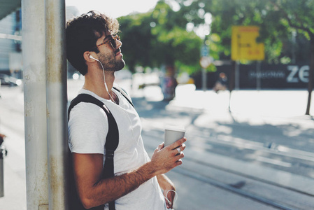 在阳光明媚的日子, 戴着墨镜的年轻人带着一杯咖啡站在大街上微笑着。大胡子男在车站等车