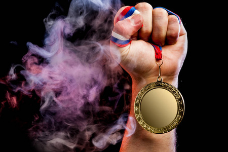 在彩色粉红色烟雾和黑色隔离背景背景下, 一只强壮的男性手拿着一枚金牌为体育成就而特写。Mocap 奖牌