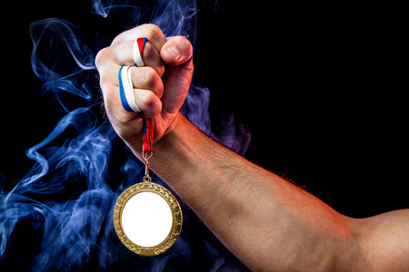 在彩色蓝色烟雾和黑色隔离背景背景下, 一只强壮的男性手拿着一枚金牌为体育成就而特写。Mocap 奖牌