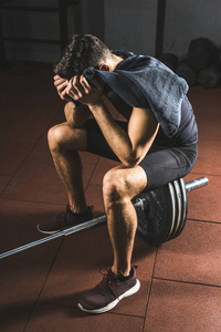 沮丧年轻运动员用毛巾在手坐在杠铃在健身房